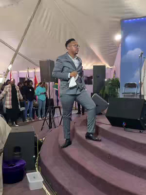 A man in a gray suit steps onto a stage in a church setting. The stage has steps and a purple carpet leading up to the front. There are speakers and people visible in the background, under a white tent-like structure.