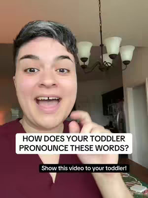 A selfie-style video featuring a woman with short, gray-streaked hair, speaking directly to the camera with an open mouth in apparent excitement. Behind her, there's a blurry view of a room with objects like a chandelier and cabinets. The woman is wearing a maroon shirt, and a text overlay is positioned at the bottom, asking viewers to show the video to their toddlers.