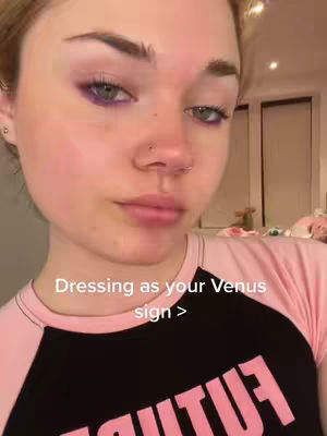 A close-up selfie of a young person with fair skin and light-colored hair. They are positioned slightly off-center, with their face angled towards the camera. The person is wearing a pink and black shirt. The text overlay reads, 'Dressing as your Venus sign >'.