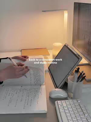 A desk setup with a computer monitor, tablet, keyboard, mouse, and writing materials. A hand holds a pen over an open notebook. Text overlayed on the image reads "back to a consistent work and study routine". The scene is lit with warm artificial light. The overall aesthetic is minimalist and clean.