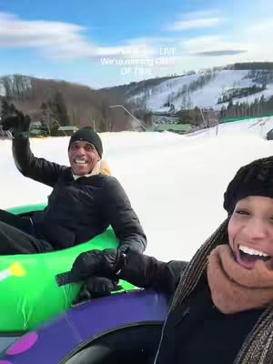 A selfie-style shot featuring a joyful couple enjoying a winter activity. A man and woman are seated in inflatable snow tubes, smiling broadly. Behind them, there's a snowy ski slope with trees and a mountain in the background under a blue sky.