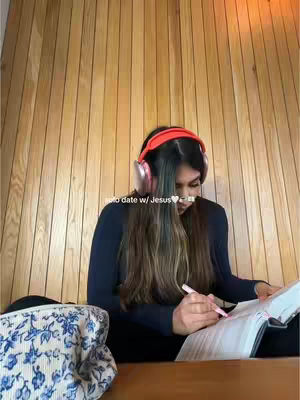 A woman with long brown hair wearing red over-ear headphones and a dark blue top is seated at a wooden table. Her hands are holding a pen and open book, while behind her is a wooden slatted wall. A patterned bag is in view. A white text overlay reads 'solo date w/ Jesus'.