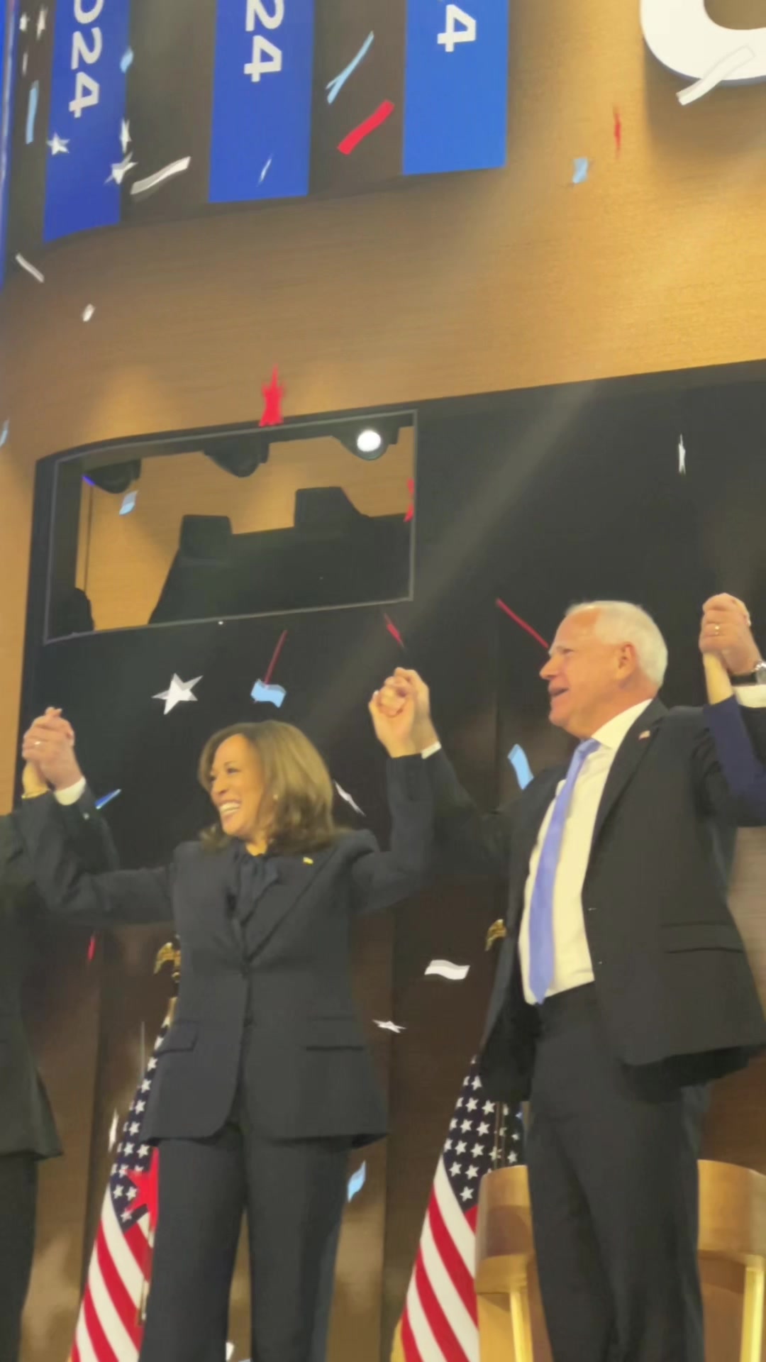 The image captures a celebratory moment with two figures holding hands and raising them in the air. In the background, there's an audience with flags with the text "2024". Confetti is falling from above. The lighting appears to be from overhead sources on a stage.