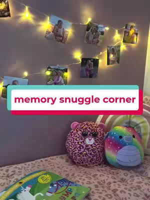 The image shows a cozy corner with a light-colored wall decorated with fairy lights and printed photos. Below the photos, there is a bed with two stuffed animals. A banner with the text, 'memory snuggle corner' is added to the lower section with a white-and-red stroke.