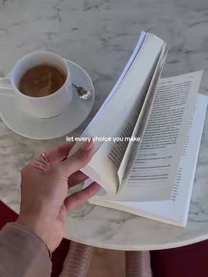 The image shows a close-up of a hand holding open a book, with a cup of coffee and saucer in the background. The book's pages are being turned, and text appears on the pages on the right side, with a cup of coffee and saucer next to it on the left side. The scene is on an off-white circular table.