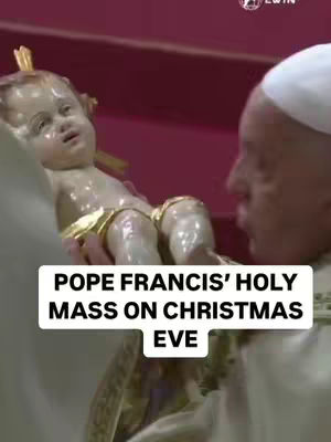 The image shows Pope Francis holding a small statue of baby Jesus with a neutral expression. The background appears to be a church setting, possibly during a mass or service.