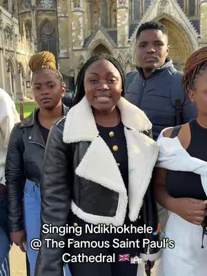 A group of people, mostly young women, stand in front of a large stone building with arched windows and columns. The central figure, a young woman, smiles and wears a black jacket with a fluffy white collar. Text overlays on the image identify the location and song.