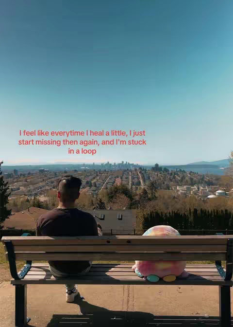 The cover image features a man sitting on a park bench, viewed from behind. He faces away from the camera, looking out over a scenic vista of trees, buildings, and a body of water. Beside him sits a plush, colorful toy. The composition is framed by a bright blue sky.
