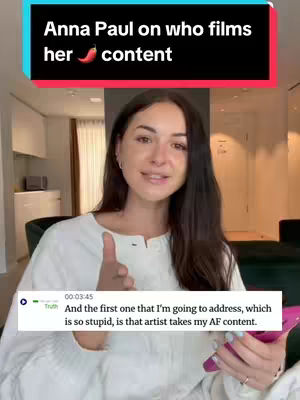 The image shows a woman, likely in her home, speaking. A black overlay at the top has white text. A text box at the bottom shows text as well. The woman on camera is a fair skinned, with dark hair, with a neutral expression, slightly smiling. She is wearing a white long sleeve top. Her right hand is raised with finger pointing. The background is a room with a white wall, natural light filtering in. The overall composition is clean and focuses on the speaker.
