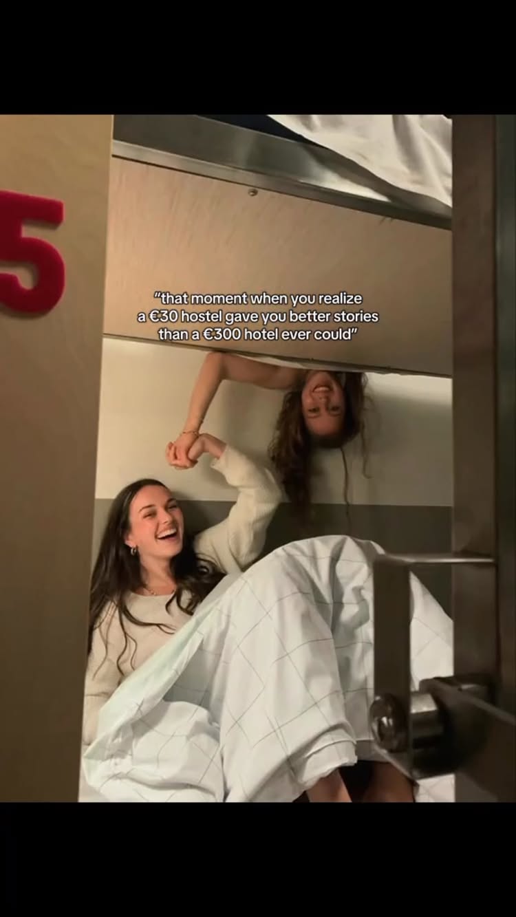 Two young women are in a hostel bunk bed. One is lying down on the bottom bunk, smiling and holding the other's hand as she leans over the top bunk. Text overlayed reads, "that moment when you realize a €30 hostel gave you better stories than a €300 hotel ever could".