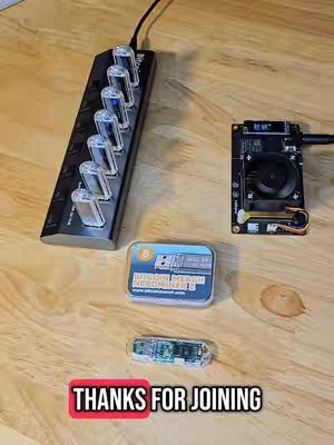 A top-down shot shows a wooden surface with electronic devices, including a black circuit board and a box with the words "Bitcoin Merch Nerdminer 2" in blue. Below is another piece of equipment, and a red rectangular graphic with the phrase "THANKS FOR JOINING" and white text.