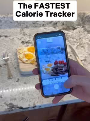 A person holds a smartphone, displaying a food image with a calorie-tracking app, in a kitchen setting. Fried eggs and strawberries sit on a small cutting board beside the phone. The background shows a marble countertop with a fork and knife.