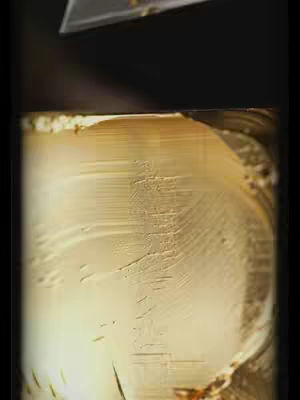 The image shows a close-up vertical shot of a golden, textured surface with a partially visible object at the top. The surface appears to be a waxy or metallic material with a subtle gradient from light to dark. The texture is detailed with fine lines and imperfections, and the lighting creates highlights and shadows that emphasize the curves and contours of the shape. The top element, possibly the edge of a tool or object, is darker and seems to interact with the shiny surface below.