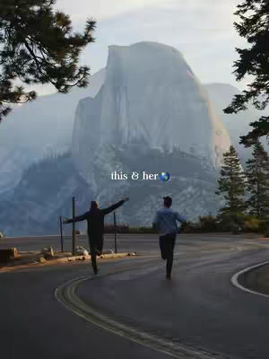 A scenic image captures two individuals running down a winding road. In the background, a towering mountain range is shrouded in a misty atmosphere. The foreground features the road, which curves invitingly, and elements of foliage that frame the scene. The overall tones are soft, with muted greens, blues, and browns. The composition feels expansive and suggests a sense of freedom and adventure.