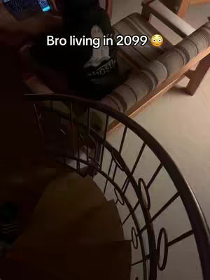 A person wearing a hoodie is seated, with a spiral staircase in the foreground. Text "Bro living in 2099" is overlayed on the image. The colors are muted, with a focus on the interaction between the person and the setting.
