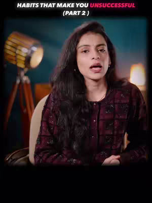 A woman with long dark hair is looking at the camera and speaking. Behind her is a lamp and a dark teal wall. The text overlay says "HABITS THAT MAKE YOU UNSUCCESSFUL (PART 2)".