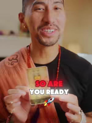 The image shows a smiling man holding a tarot card toward the camera, with overlaid text reading 'So are you ready'. A rocket ship graphic adds an interactive feel to the image. The man presents the card in a brightly lit setting, with his face centered and well-lit.