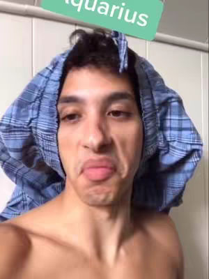 A selfie-style shot featuring a light-skinned man with dark hair. He is shirtless and has a towel wrapped around his head. A green rectangular label with the word 'Aquarius' in white text is positioned above his head. The man has a slight grimace and his lips are pursed. The background is a neutral white wall.