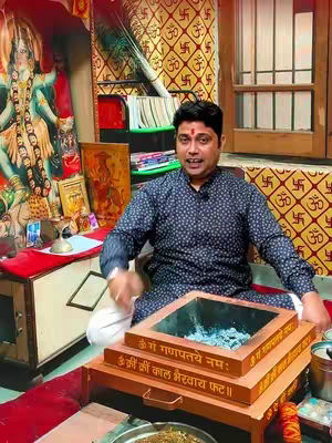 A man in a dark blue patterned shirt with a red bindi sits in front of a traditional setup with a fire place. The background features religious motifs and symbols.