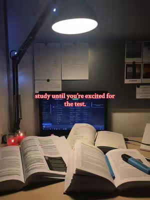 The image is an indoor shot of a desk setup for studying at night. In the foreground, several open books and a highlighter are visible, suggesting active study. Above the books, a computer screen is visible displaying content. A desk lamp with a large, circular light is positioned above, illuminating the desk. A black wall is in the background. The overall color scheme is dark, with warm light from the lamp and candles, creating a focused study atmosphere. Text overlayed in the image reads 'study until you're excited for the test'