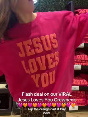 A woman is shown from the chest up, wearing a bright pink sweatshirt with large, orange text that reads "JESUS LOVES YOU". The background appears to be a retail setting, with a glimpse of shelves stocked with similar sweatshirts. Text overlays at the bottom describe a flash deal, the product being the "VIRAL Jesus Loves You Crewneck," and prompts viewers to tap an orange cart to buy now, accompanied by heart emojis.