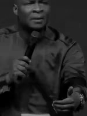 A monochrome image of a man speaking into a microphone, likely in a public setting. The man, dressed in a dark-colored jacket, is captured from the mid-chest and up, with the microphone held near his mouth. His right hand and forearm are visible, with a wrist watch.