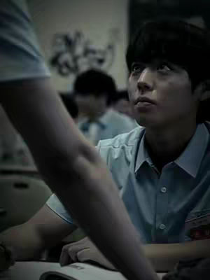 A medium shot shows a young man in a light blue button-down shirt looking up and slightly to the right. His dark hair is neatly styled. In the foreground, a blurred arm partially obscures the left side. The background is out of focus, suggesting a school setting or similar indoor environment. The lighting is dim, with muted tones and a slightly desaturated color palette, creating a somewhat dramatic or somber mood.