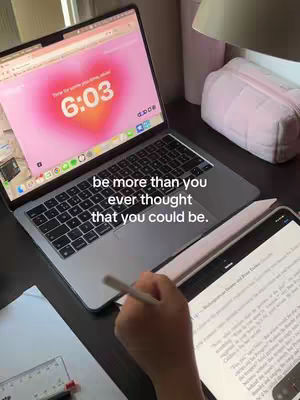 The image shows a laptop and a tablet on a desk. The laptop screen displays a pink heart-shaped background with the time 6:03. The tablet has a document with text. There is a text overlay at the bottom center of the image that reads: "be more than you ever thought that you could be." The hand holding a stylus for the tablet can also be seen.