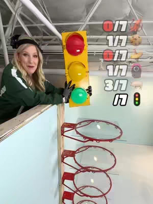 A woman in a green outfit stands on a wooden ledge, holding a prop traffic light with a red light on, ready to drop objects into basketball hoops beneath. Score indicators sit on the right side of the image.