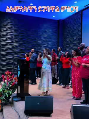 A live performance takes place on a stage. A woman in a light-colored dress holds a microphone and sings. Behind her, a group of people, some holding microphones, sing along. The background features a dark textured wall with blue lighting. The foreground includes speakers and a floral arrangement. The lighting is bright and focused on the performers.