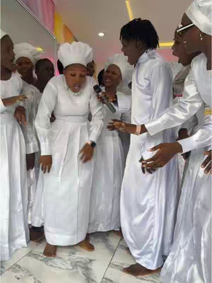 A group of people in white robes are gathered indoors. The lighting is bright and appears to be in a sanctuary or similar setting. There are multiple figures, all dressed in white, some with head coverings. The central focus is on a person who is holding a microphone and seems to be leading or performing, with the rest of the group engaging actively. The overall composition suggests a religious or spiritual performance.