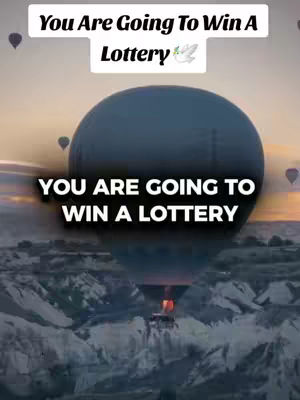 The image showcases a dramatic landscape with a large hot air balloon dominating the center. The balloon is dark blue or black, and its shape is a prominent visual element. Below the balloon, rugged mountains and a valley create a textured backdrop. Overlaid text in white capital letters announces 'YOU ARE GOING TO WIN A LOTTERY'. Colors appear to transition from darker hues to a vibrant sunrise, creating a visually appealing aesthetic, and there are a few more balloons in the sky.
