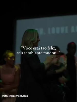 A woman with blonde hair is speaking on a stage, likely at a church. Text overlays on the image. The woman is wearing black clothing, and the background is out of focus, with other people visible. The text overlay reads, “Você está tão feliz, seu semblante mudou..”, which translates to “You are so happy, your face has changed..”, suggestive of a personal transformation conveyed through faith.