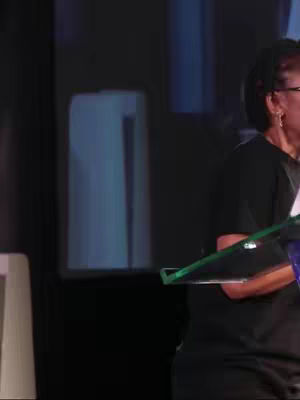 A woman with dark hair and glasses stands on a stage, holding a clear award with a stage background. The image is composed primarily of blacks, blues, and greens.