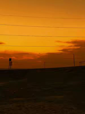 A silhouetted landscape at dusk or dawn. The sky is predominantly orange and yellow with some cloud cover along the horizon. Silhouetted shapes suggest land, some posts, and the barest hint of structures.