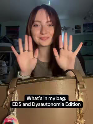 A young woman with long auburn hair is smiling and holding her hands up with all fingers spread wide, displaying several rings. Behind her, there is a Coach tote bag with the text overlayed in the bottom third of the image.