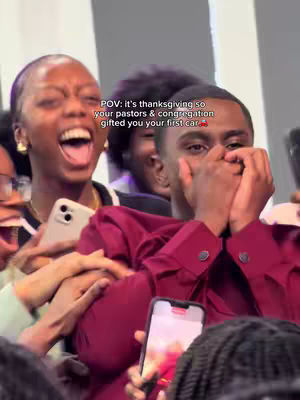 A group of people, primarily black in complexion, are gathered indoors, likely at a church or event. The focus is on a young man in a burgundy shirt who has his hands covering his face, likely in a moment of surprise or joy. A woman on the left is expressing excitement with her mouth wide open, and holding a phone. The background is blurred, but appears to be a well-lit space. The overall colors are warm, with the burgundy shirt providing a focal point. A phone recording is also visible.