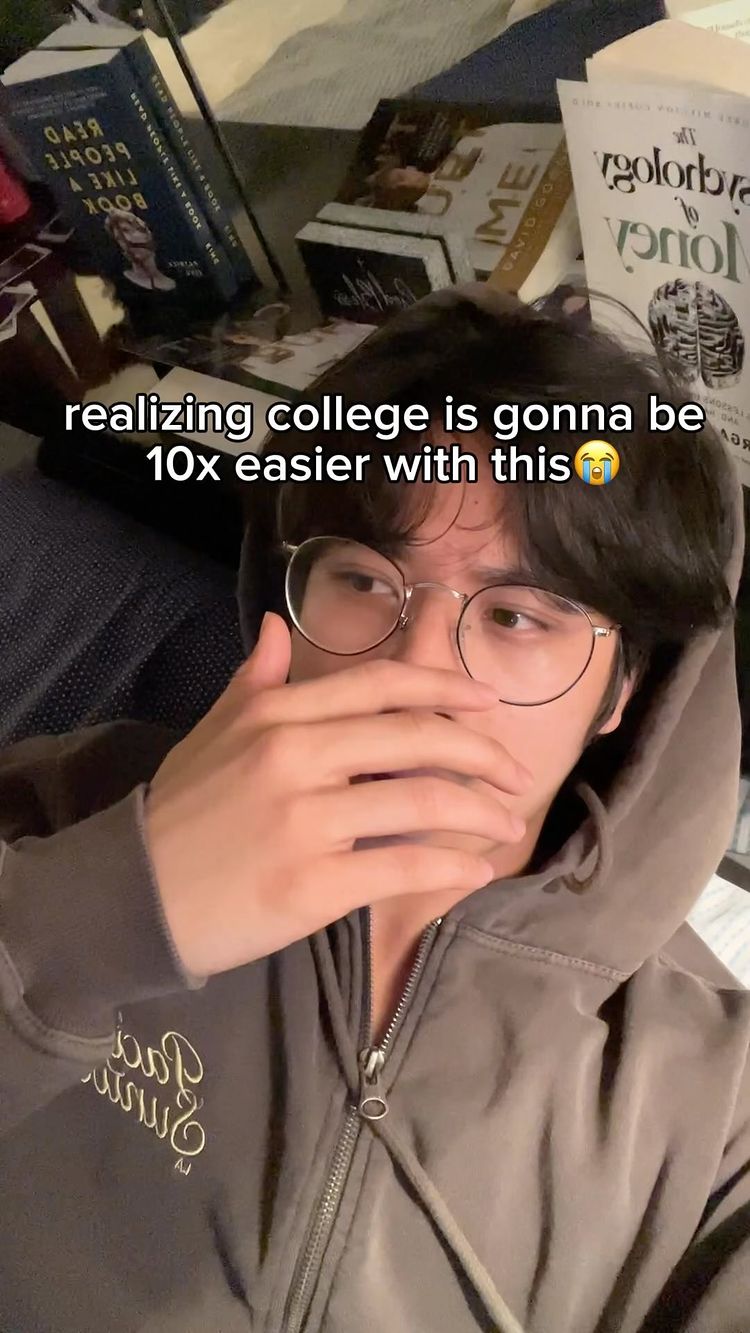 A young man with dark hair is shown close-up, covering his mouth with his hand, wearing glasses and a grey hoodie. Behind him are books and a cluttered background.