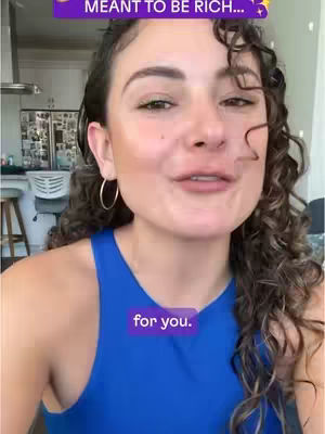 A woman with curly hair in a blue top is looking at the camera and speaking. There is text overlaid on the image in two places. In the background there is a kitchen.