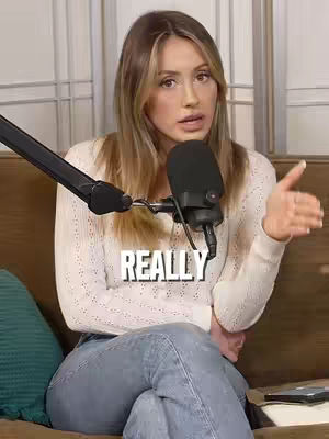 A woman with long blonde hair and a white sweater is seated and speaking into a microphone. Text overlayed on the image reads 'REALLY.' The background consists of light-colored vertical panels.
