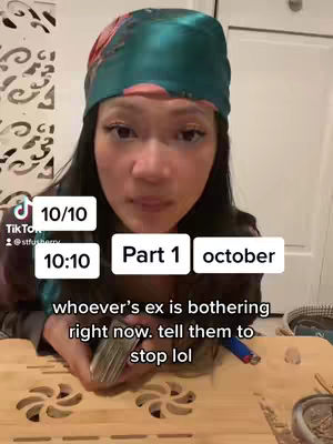 A woman with long dark hair and light skin is centered, wearing a teal patterned headscarf and dark clothing. The background is out of focus, a simple white door and patterned wall are lightly visible. Text overlays at the top left and center right provide context. She's holding a deck of cards in her hands with a table setting. The lighting seems interior, with a soft focus on the woman's face.