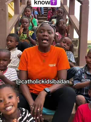 A woman in an orange shirt is surrounded by children of various ages, all seated on steps. The woman is speaking with an open mouth, and the children are looking towards the camera.