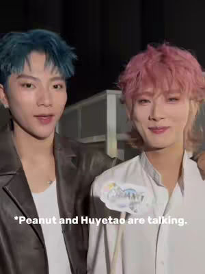 Two young men are seen in a side-by-side pose. The man on the left has blue hair and wears a leather jacket over a white shirt. The man on the right has pink hair and wears a white shirt. The background is dark.