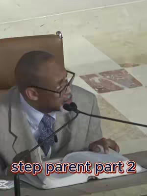 The image shows a man in the middle of a speech. He is wearing glasses, a suit, and a tie. He is holding a book and speaking into a microphone. The background is a room with a tiled floor and a patterned wall. The text overlay at the bottom says 'step parent part 2'.