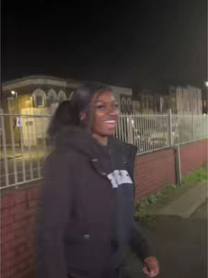 A woman is walking and smiling at night. She is wearing a black jacket over a dark hoodie. The background behind her includes a building and a fence.