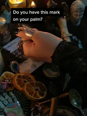 A close-up shot of a hand adorned with a silver ring holding small objects on a table with various mystical items. In the background, there are candles and what appears to be a bust of a person.