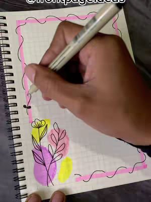 A hand holding a black pen is drawing with a highlighter on a white spiral notebook. The drawing includes a pink border, a flower, leaves, and some abstract shapes colored in pink and yellow.