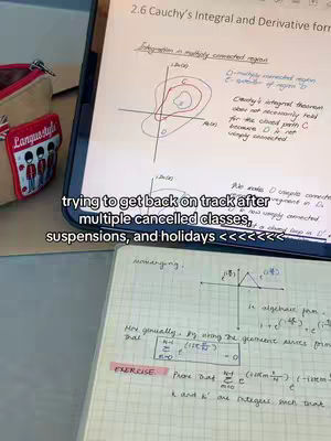 The image shows a notebook and tablet with mathematical or scientific notes. A pencil case is on the left.
