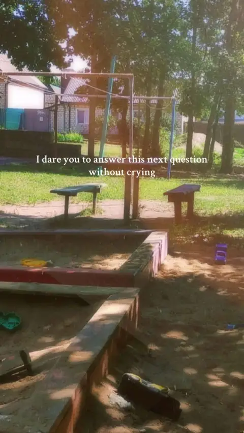 The image captures a sunny outdoor scene centered around a sandbox. The sandbox is made of rectangular brick walls. A wooden object and some toys are on the sand. There are trees, a building, and a bench in the background under the sunlight. The sky is filled with sunlight.