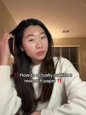 A young woman with long dark hair touches her hair and looks at the camera. The background appears to be an interior room lit by a warm light. Text overlayed says, "How to actually publish a research paper !!".